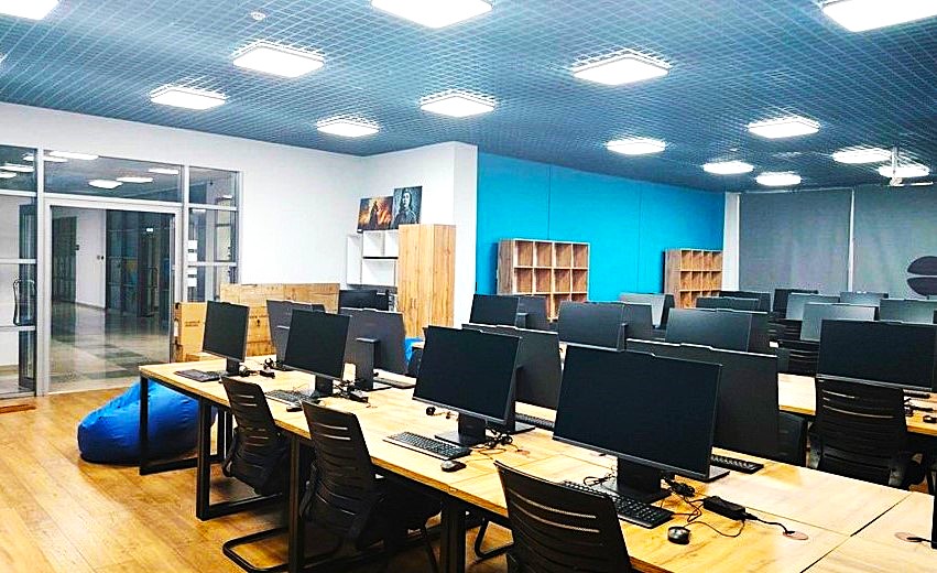 Заманауи компьютерлік сынып: ұзақ үстелдер, мониторлар мен кеңсе орындықтары орналасқан жарық бөлме.
