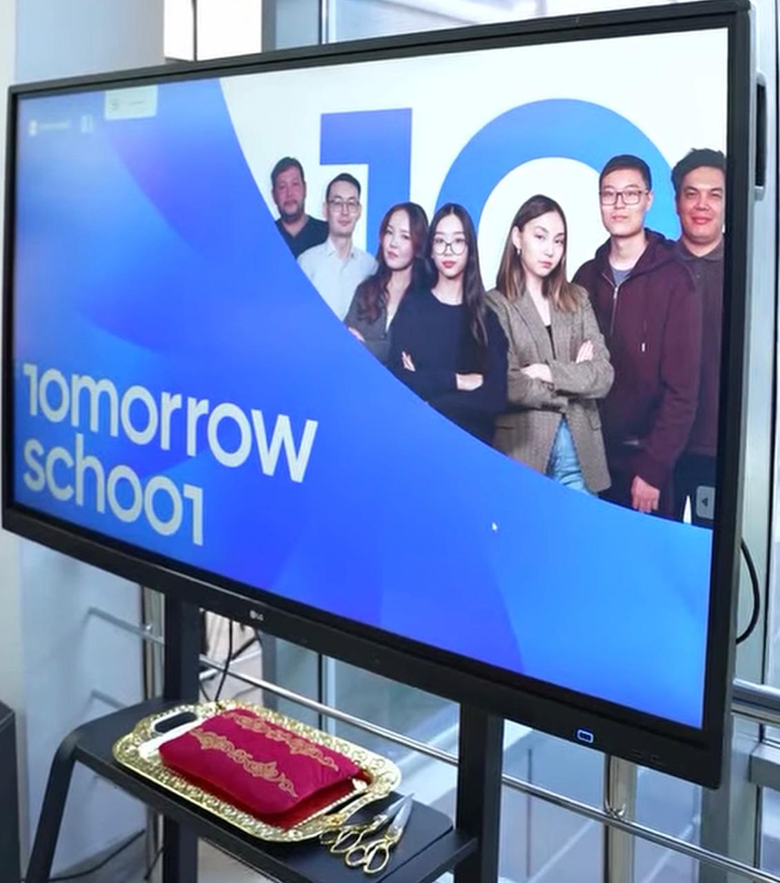 «Tomorrow School» жазуы мен адамдар бейнеленген үлкен экран және алдындағы сыйлық қоймасы.