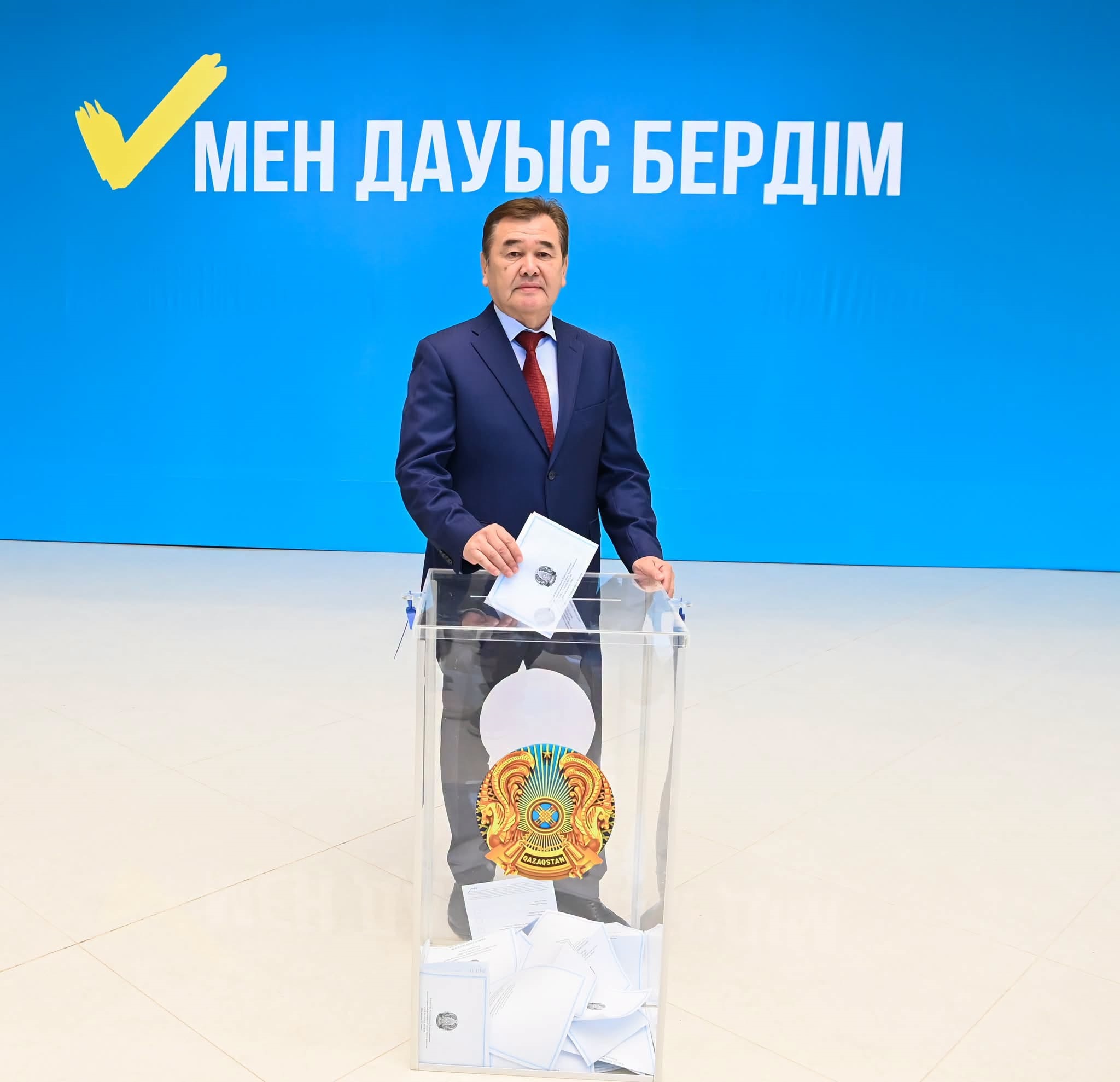 Облыс әкімі дауыс қорабына бюллетень тастап жатыр. Артында «Мен дауыс бердім» деген жазу бар.