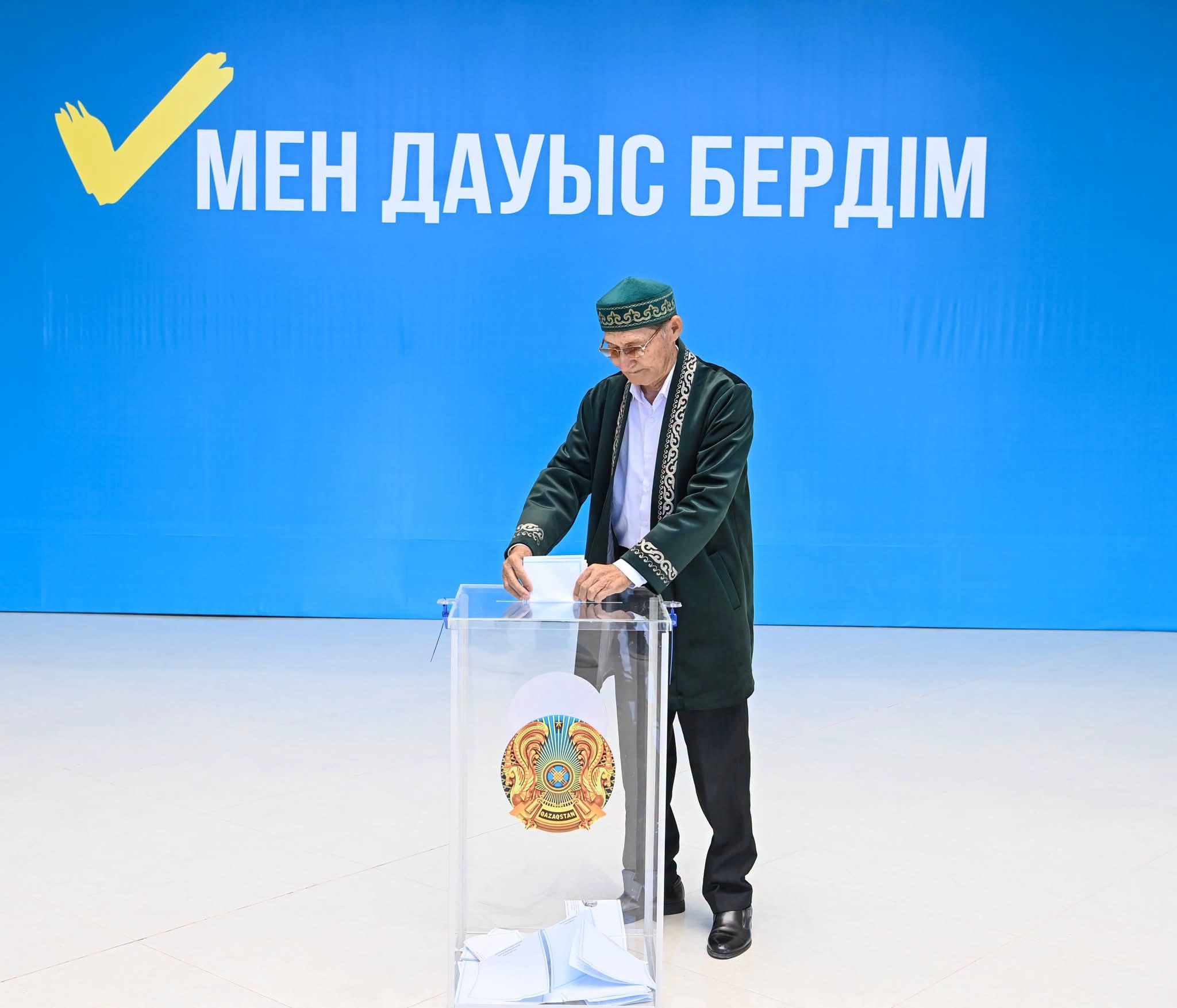 Ұлттық киім киген ер адамның дауыс қорабына бюллетень салуы. Фонда «Мен дауыс бердім» жазуы бар.