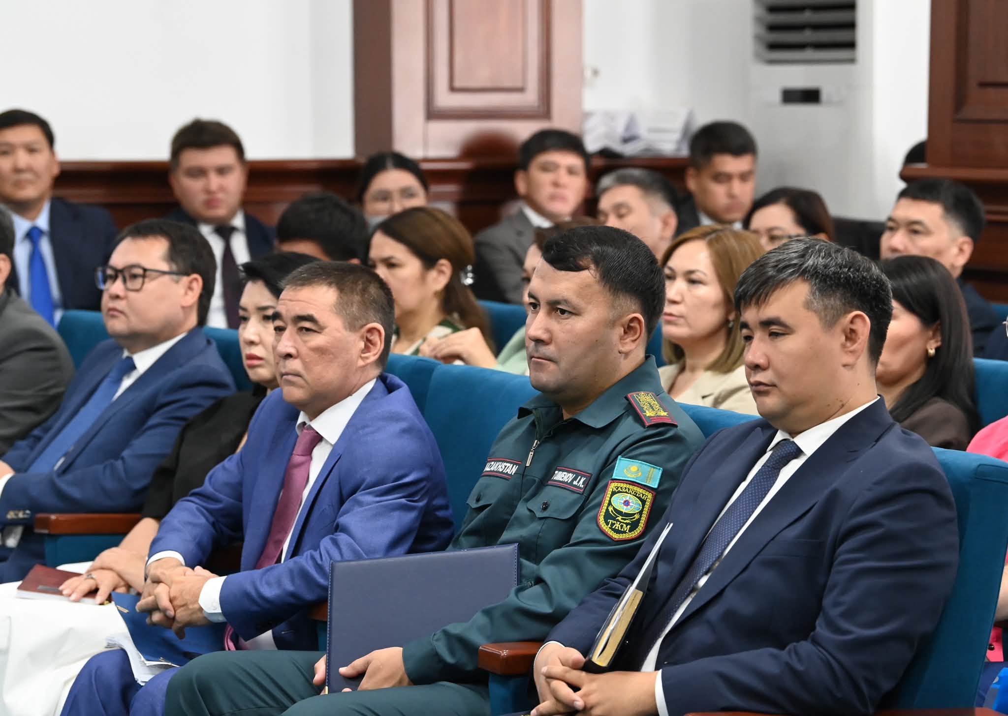 Жиналыста отырған костюм киген және әскери формадағы ерлер мен әйелдер.