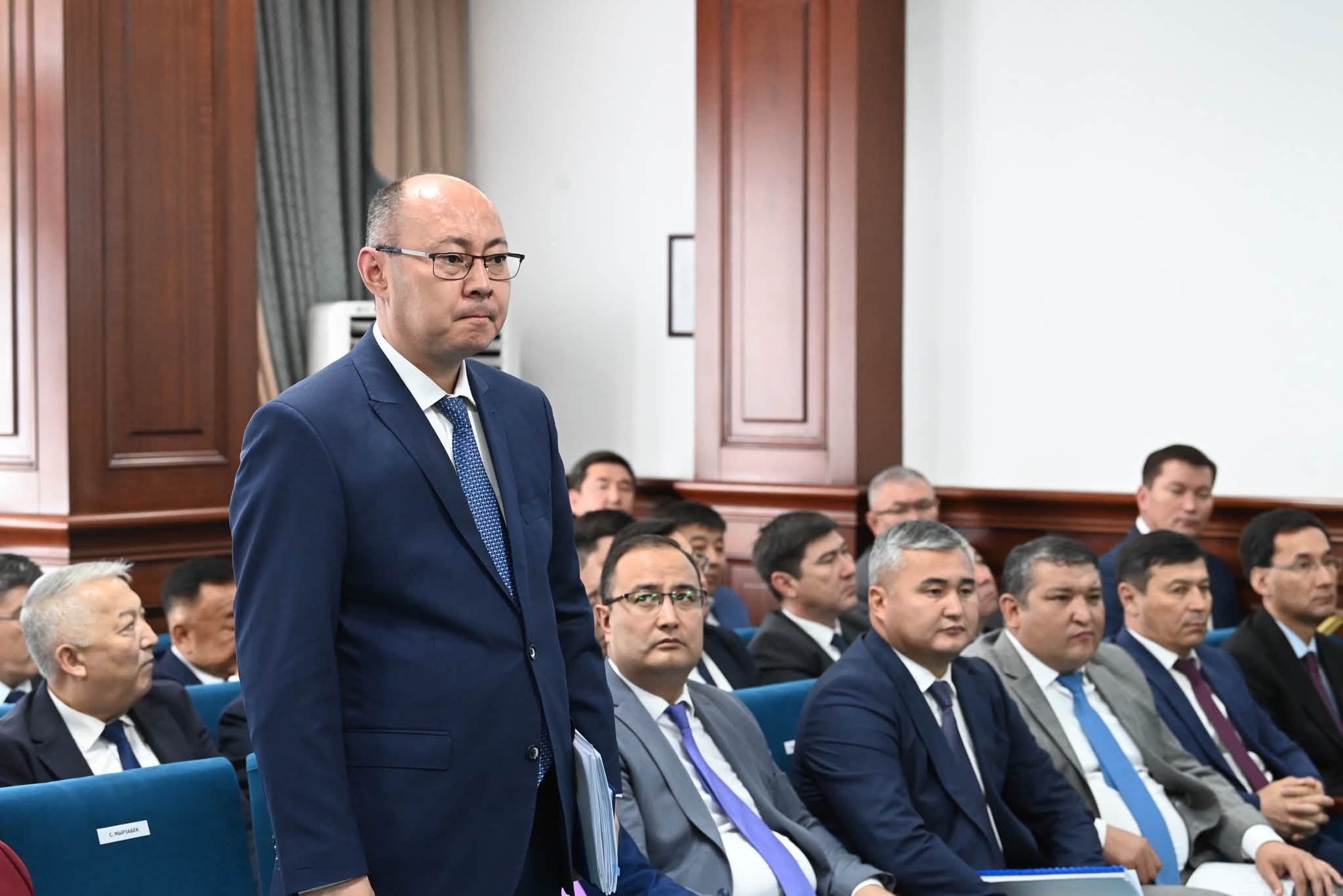 Көк костюм киген, көзілдірікті ер адам жиналыста тұрып сөйлеп жатыр.