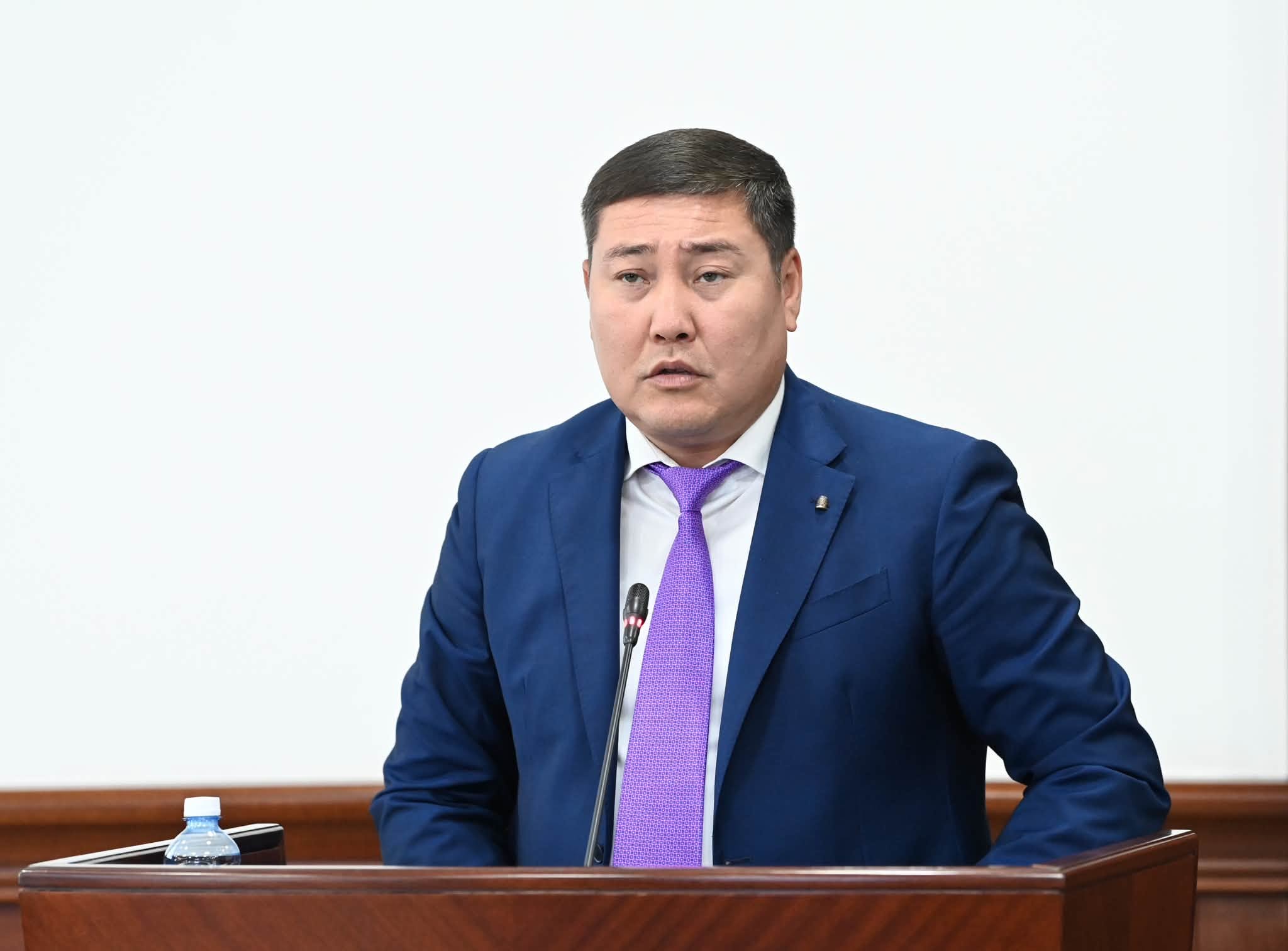 Көк костюм мен күлгін галстук киген ер адам трибунада сөйлеп тұр.