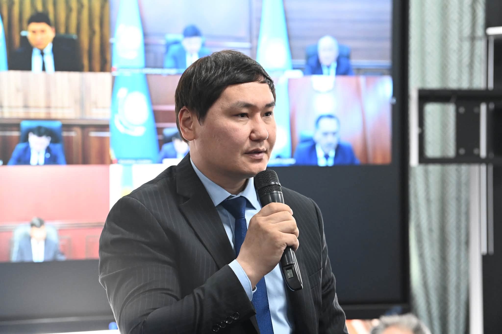 Микрофон ұстап, сөйлеп тұрған костюм киген ер адам.