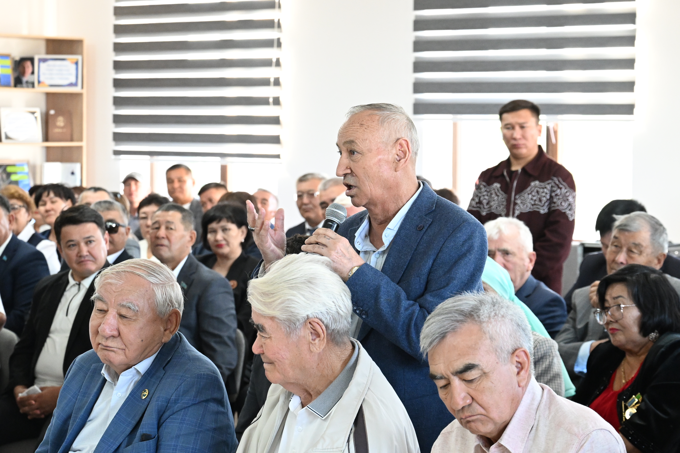 Жиынға жиналған адамдардың арасында микрофонмен сөйлеп тұрған қарт кісі.