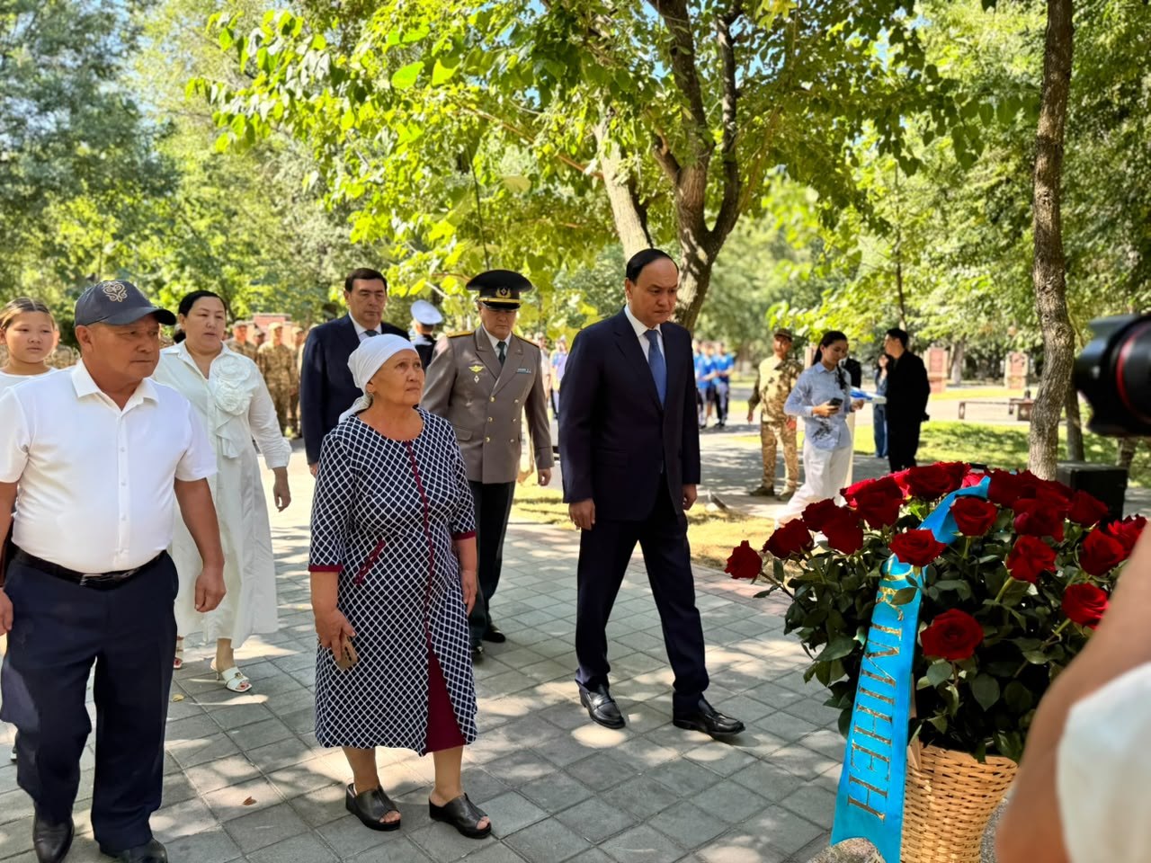 Ресми киінген адамдар мен қария әйелдің гүл шоғының алдында тұрғаны.