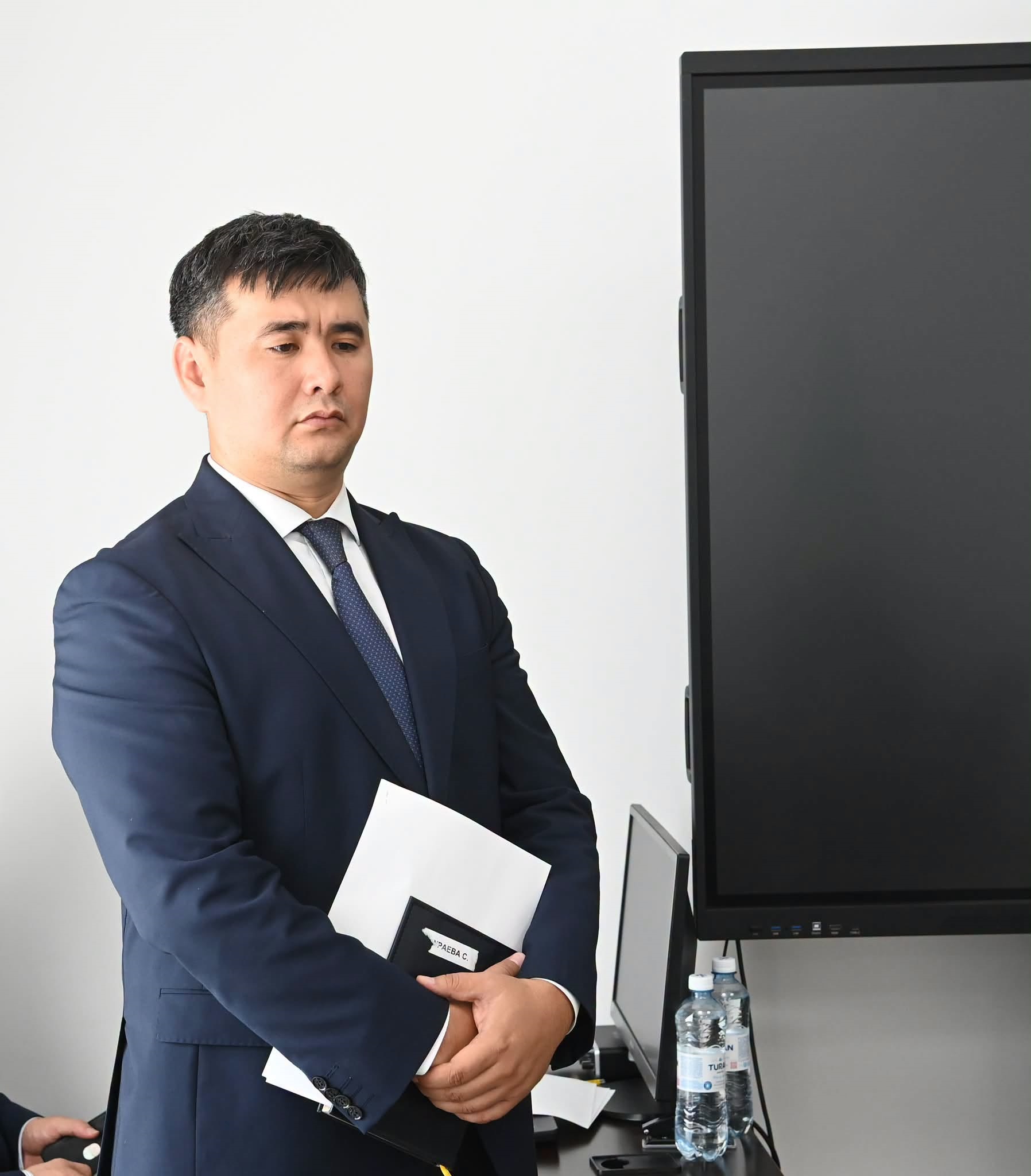Костюм киген, қолында құжаттары бар ер адам монитордың жанында тұр.