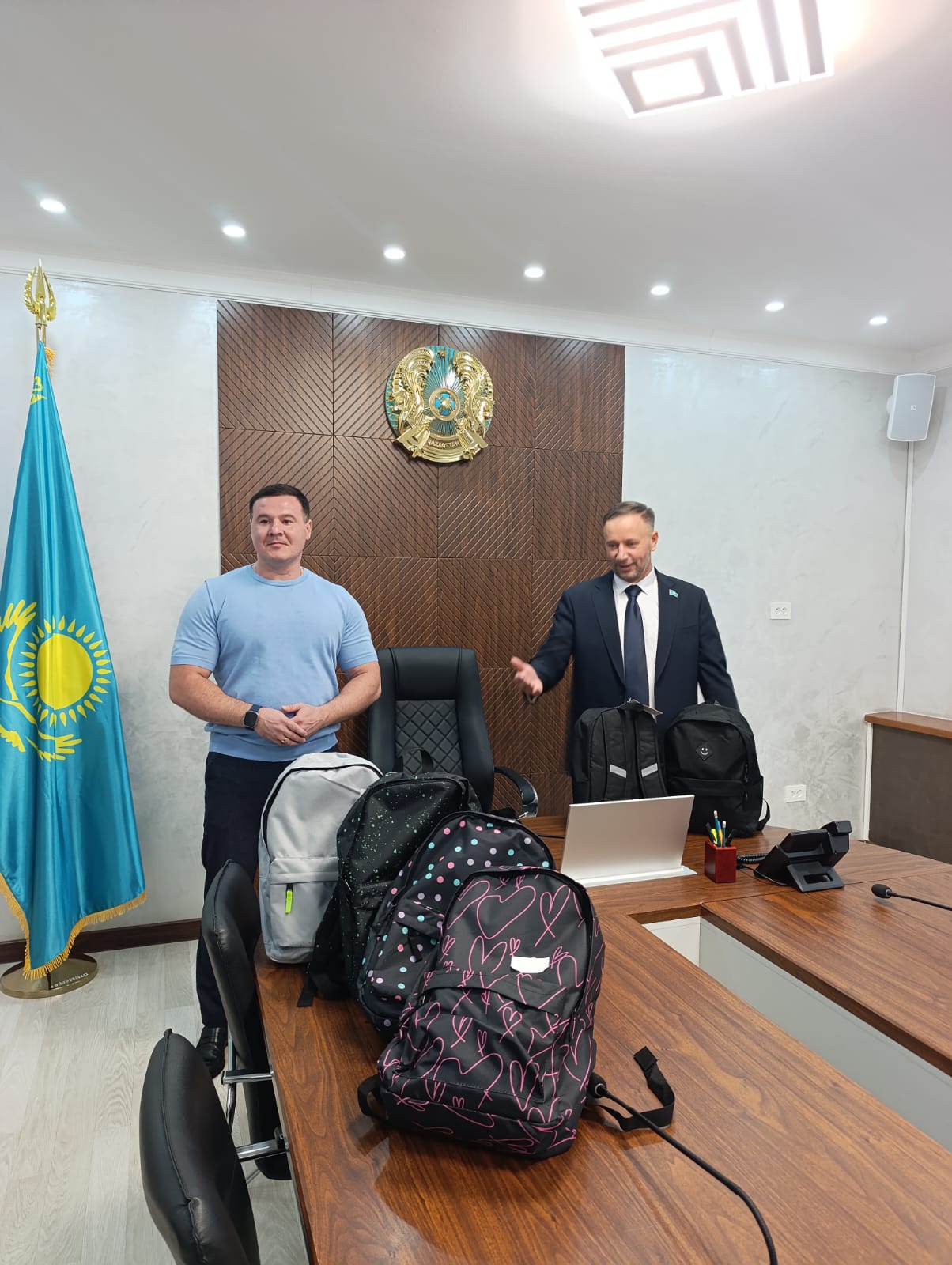 Қазақстан Республикасының туы мен елтаңбасы бейнеленген фонда кеңседе отырған екі ер адам, үстел үстінде мектеп сөмкелері тұр