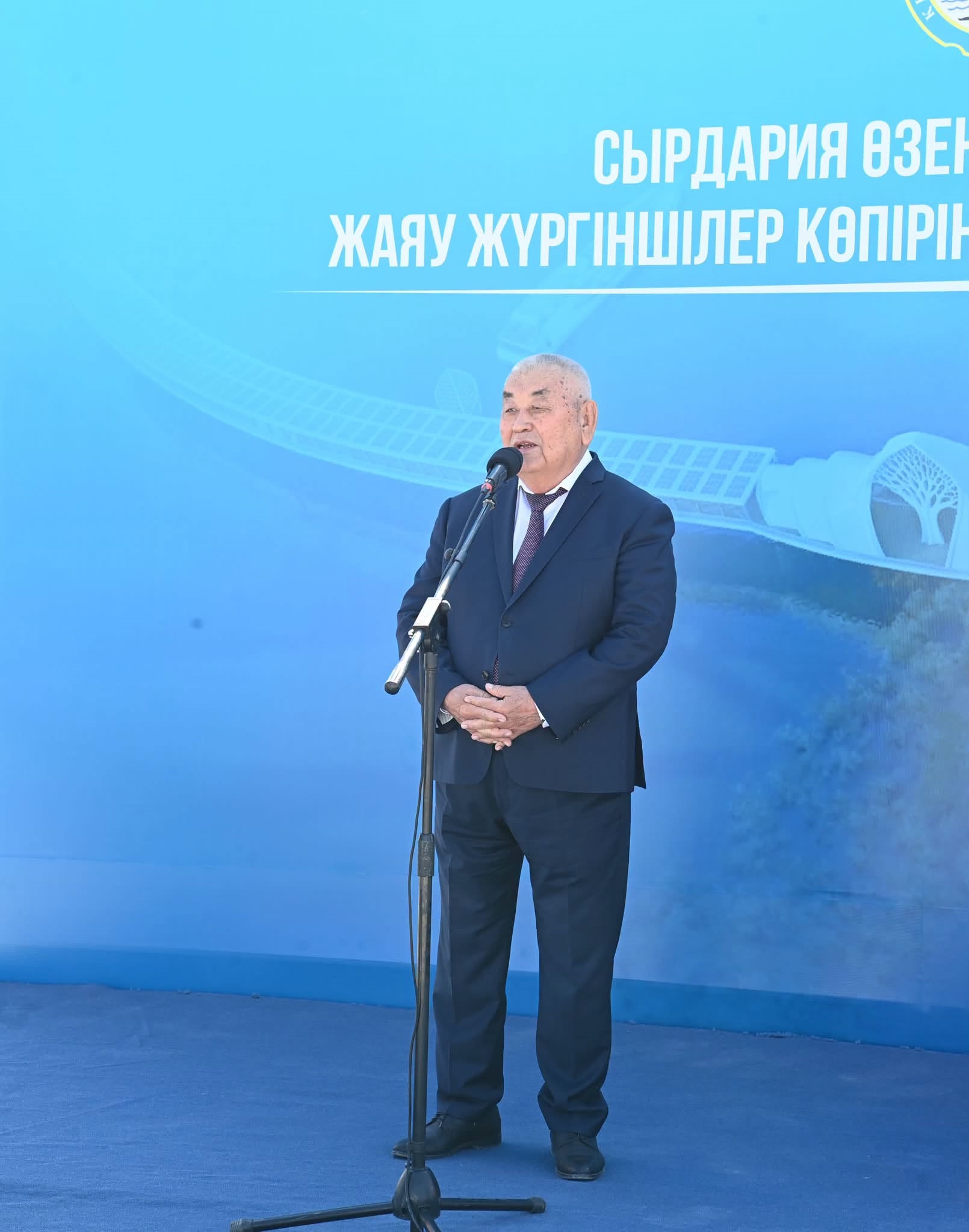 Көк түсті фон алдында микрофонмен сөйлеп тұрған костюм киген ер адам.