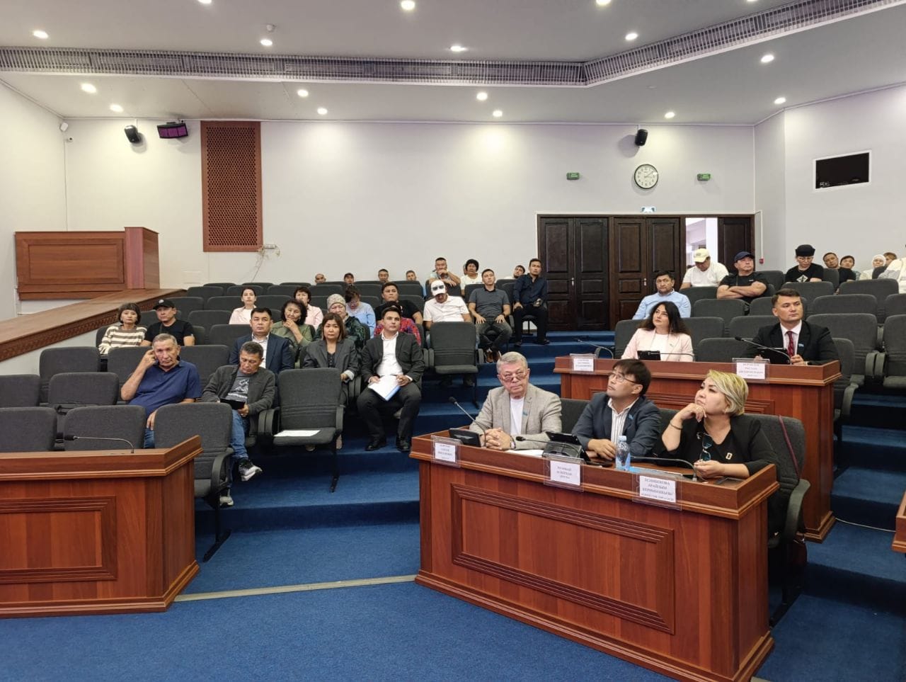 Конференц-залдағы конференцияда ағаш үстелдерде және аудиторияда отырған адамдар тобы.