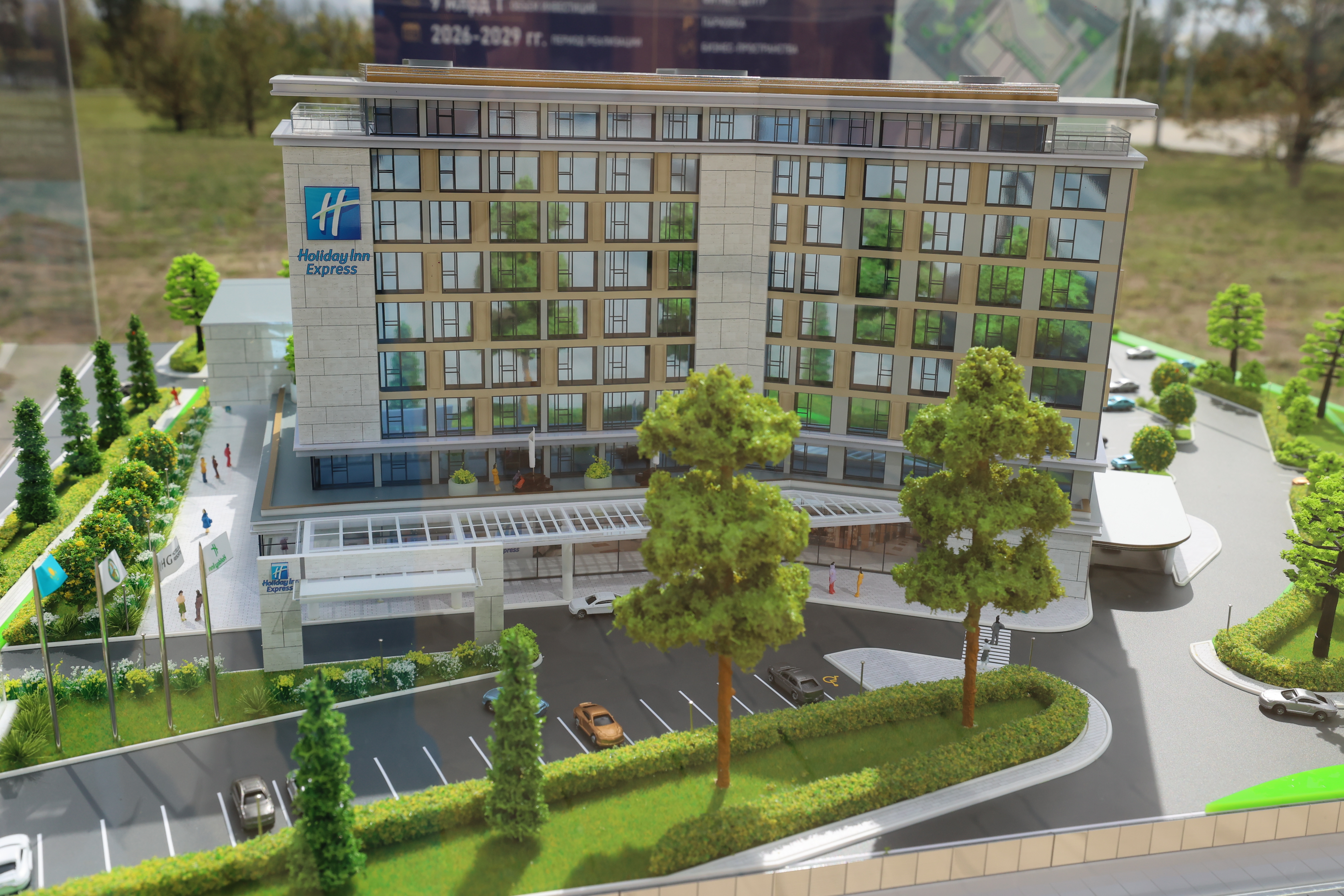 Заманауи көп қабатты Holiday Inn Express қонақ үйінің архитектуралық макеті, оның айналасы мен көгалдандыруы бар.