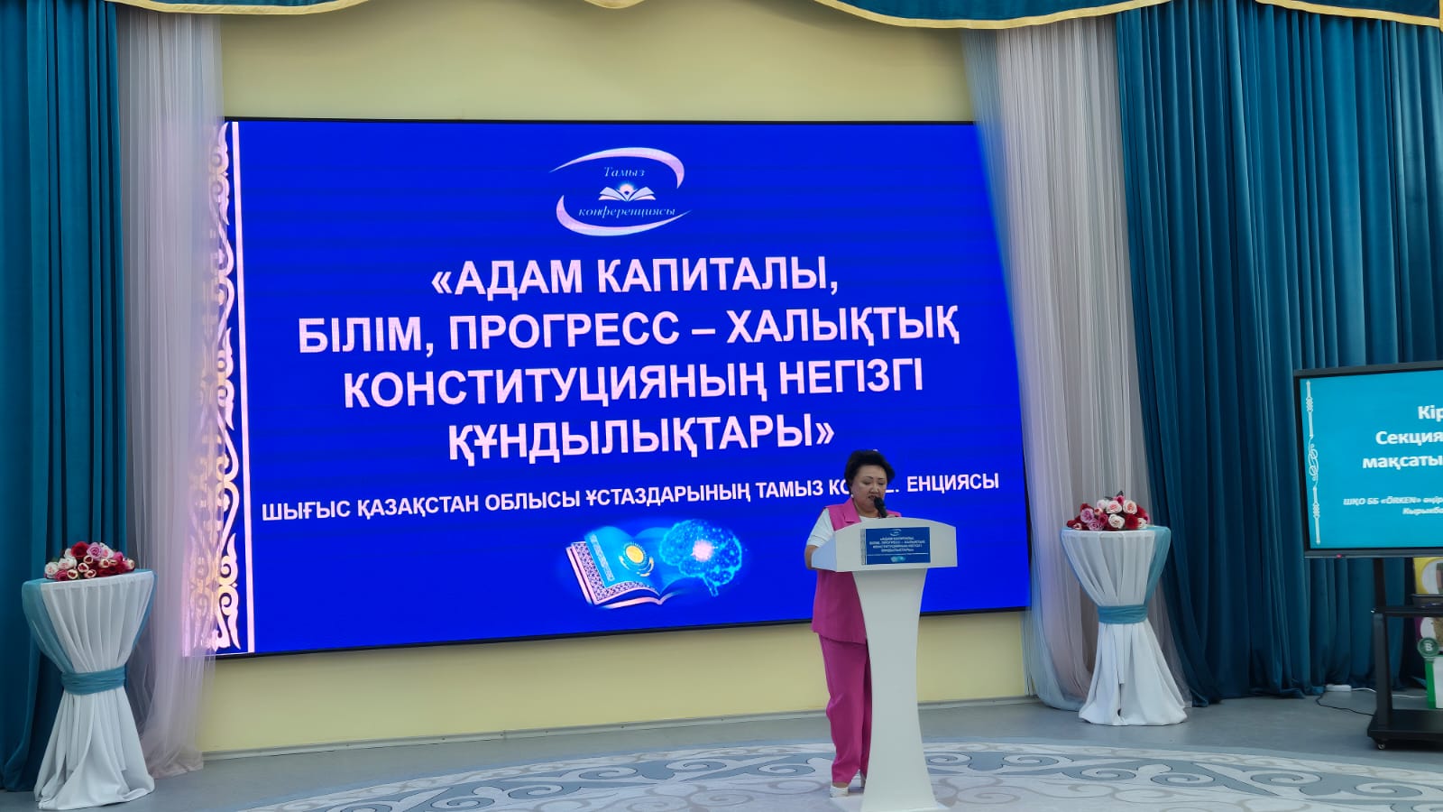 Конференцияда сөйлеп тұрған спикер және «Адам капиталы, білім, прогресс» тақырыбы жазылған үлкен экран.