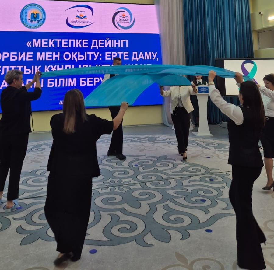Женщины в деловой одежде держат большую голубую ткань на фоне баннера конференции по дошкольному образованию.