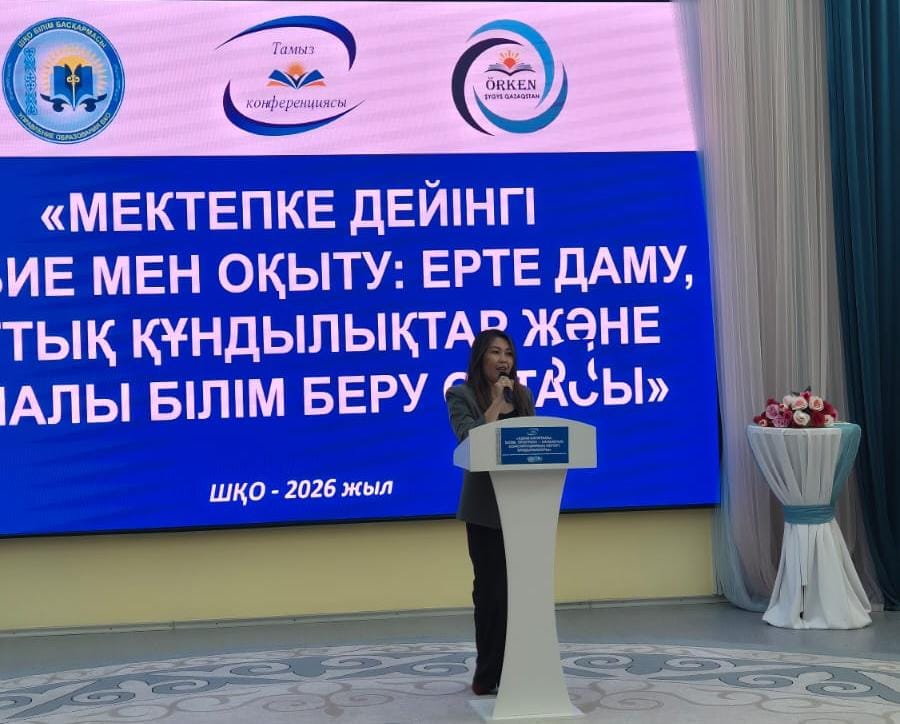 Женщина выступает с речью за трибуной на фоне большого экрана с текстом конференции на казахском языке.