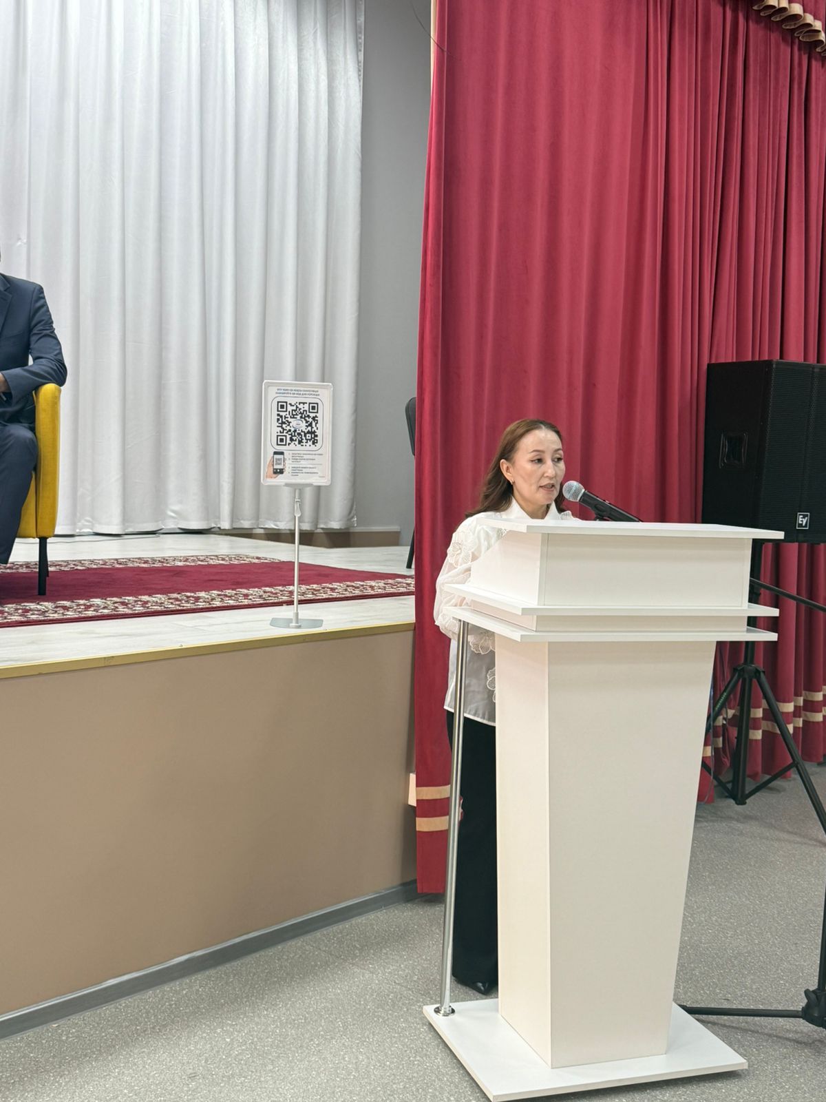 Ақ трибунада тұрып, микрофон арқылы сөйлеп жатқан әйел. Фонда қызыл перде мен ақ зана бар.