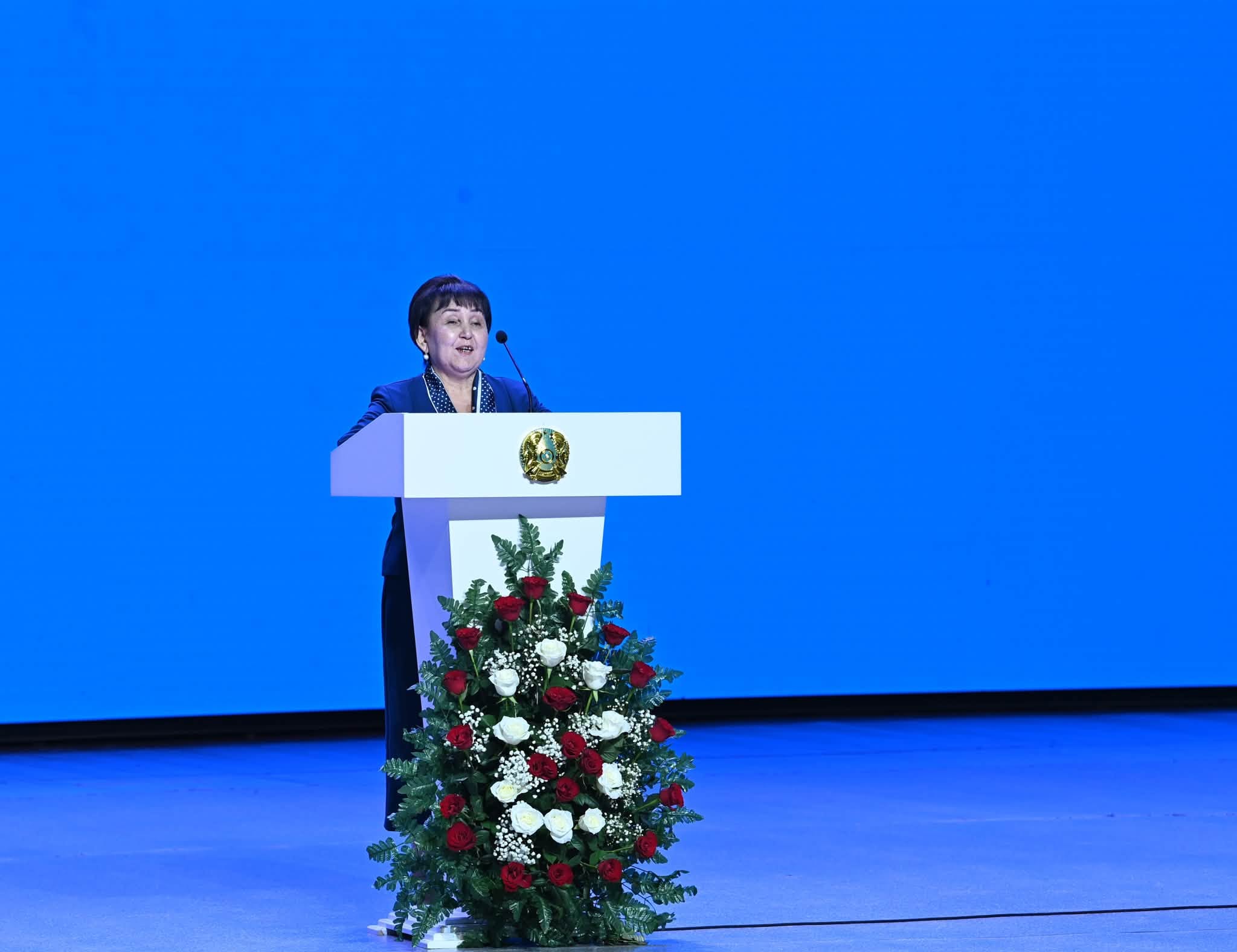 Көк фонды сахнада, гүлдермен безендірілген трибунада сөйлеп тұрған әйел.