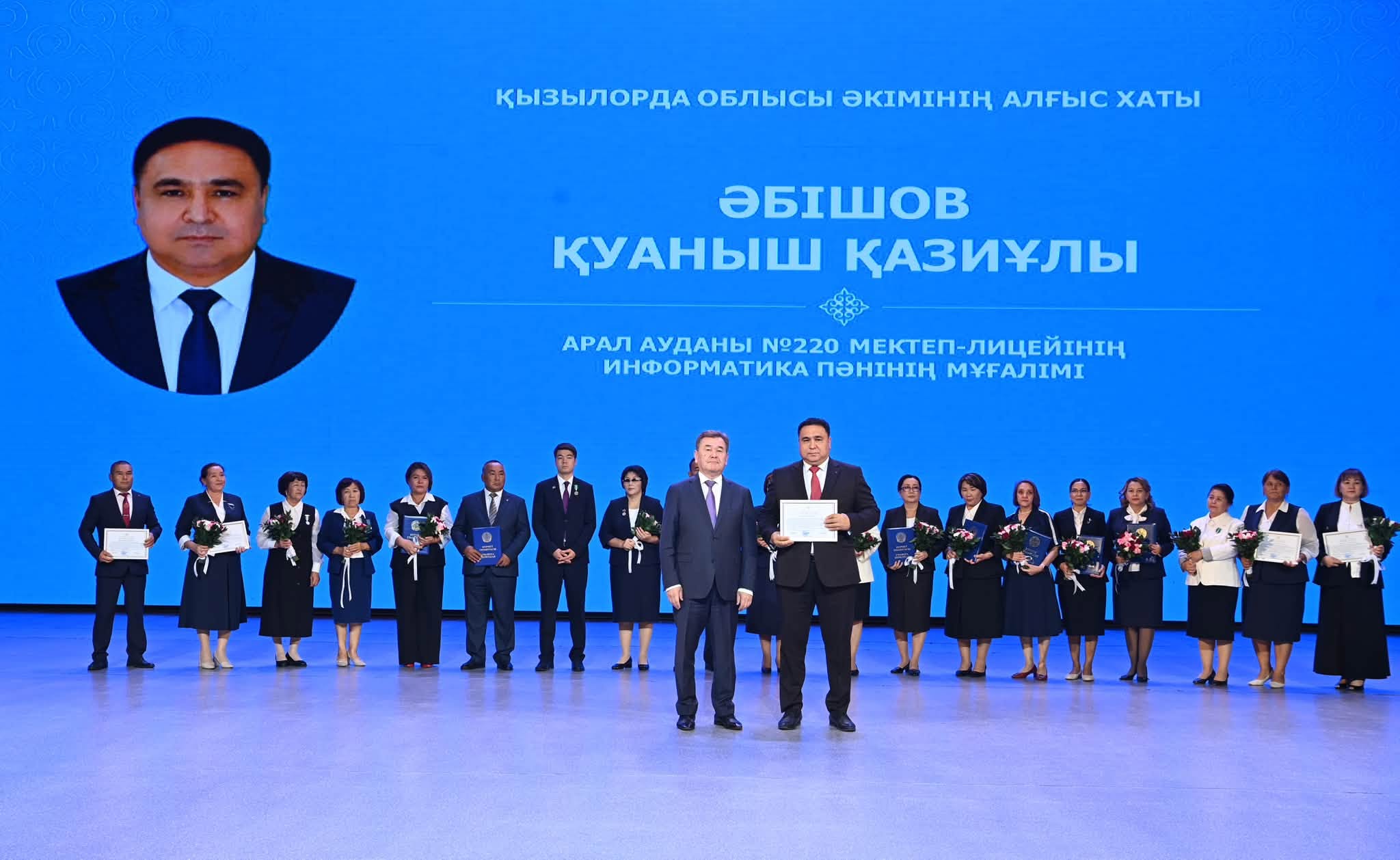Марапатталған мұғалімдер мен қонақтар сахнада. Фонда Әбішов Қуаныш Қазіұлына арналған алғыс хат бейнеленген.