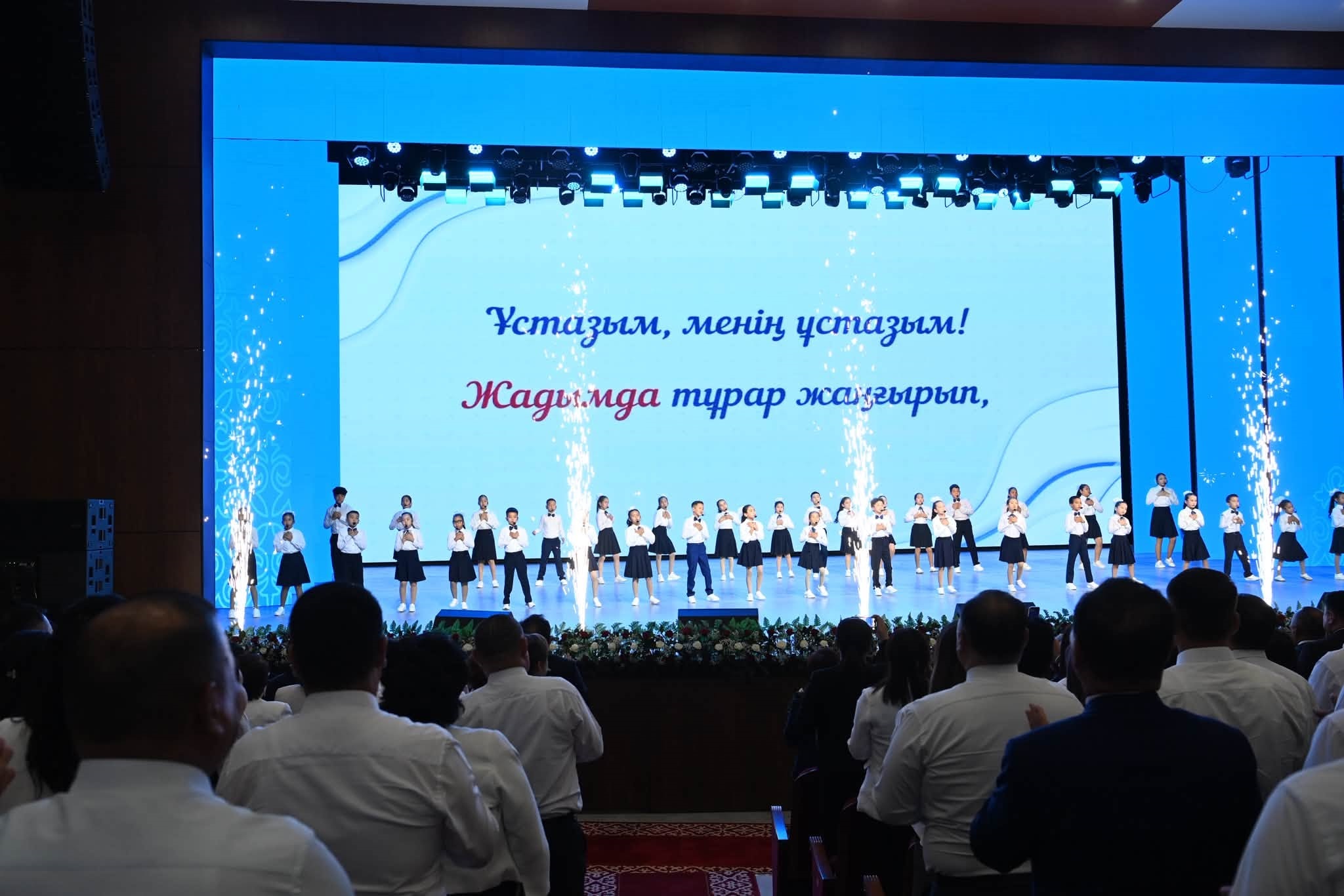 Сахнада ән айтып тұрған балалар және артқы фондағы «Ұстазым, менің ұстазым!» деген жазуы бар экран.