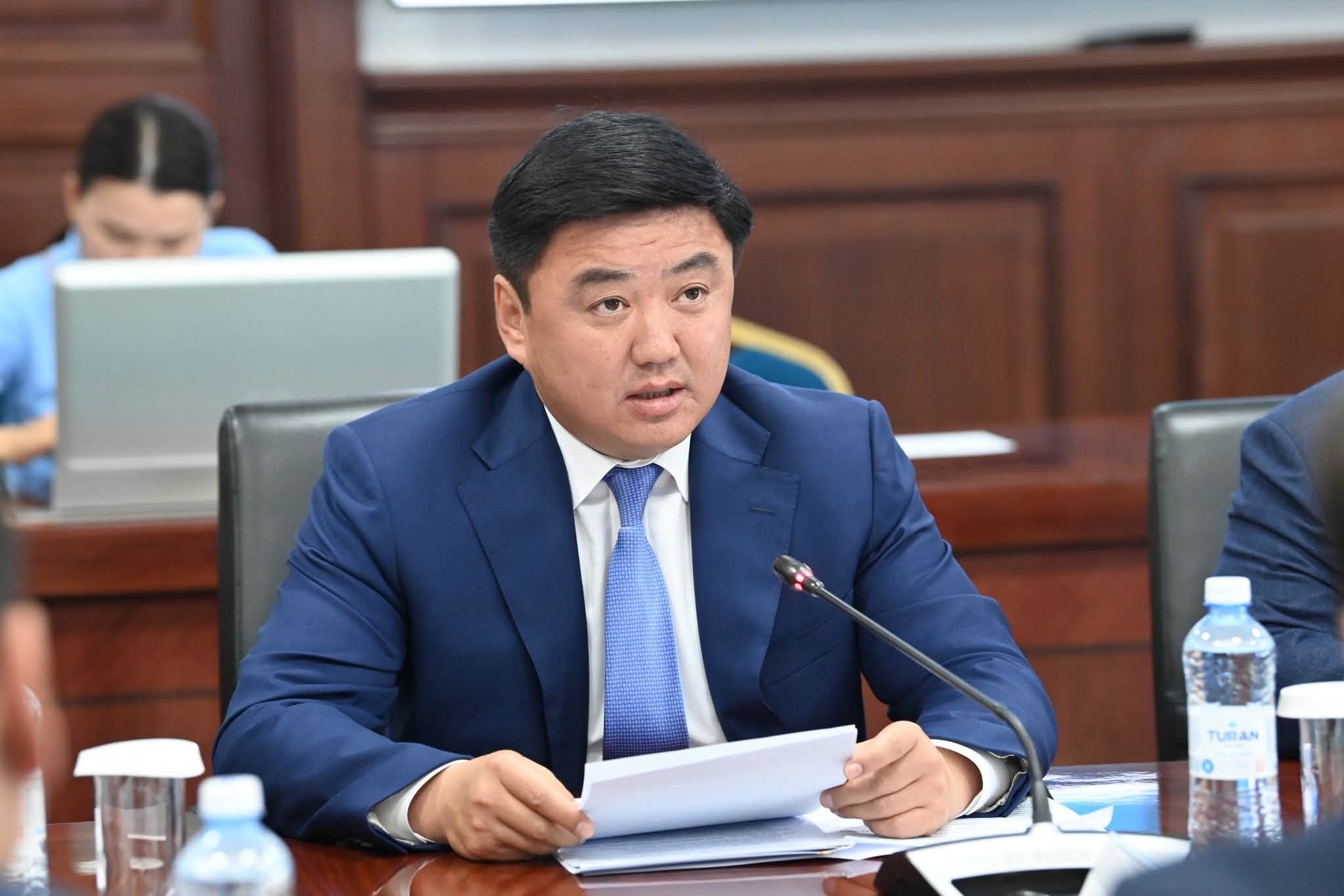 Көк костюм киген ер адам жиналыста құжат ұстап, сөйлеп тұр.