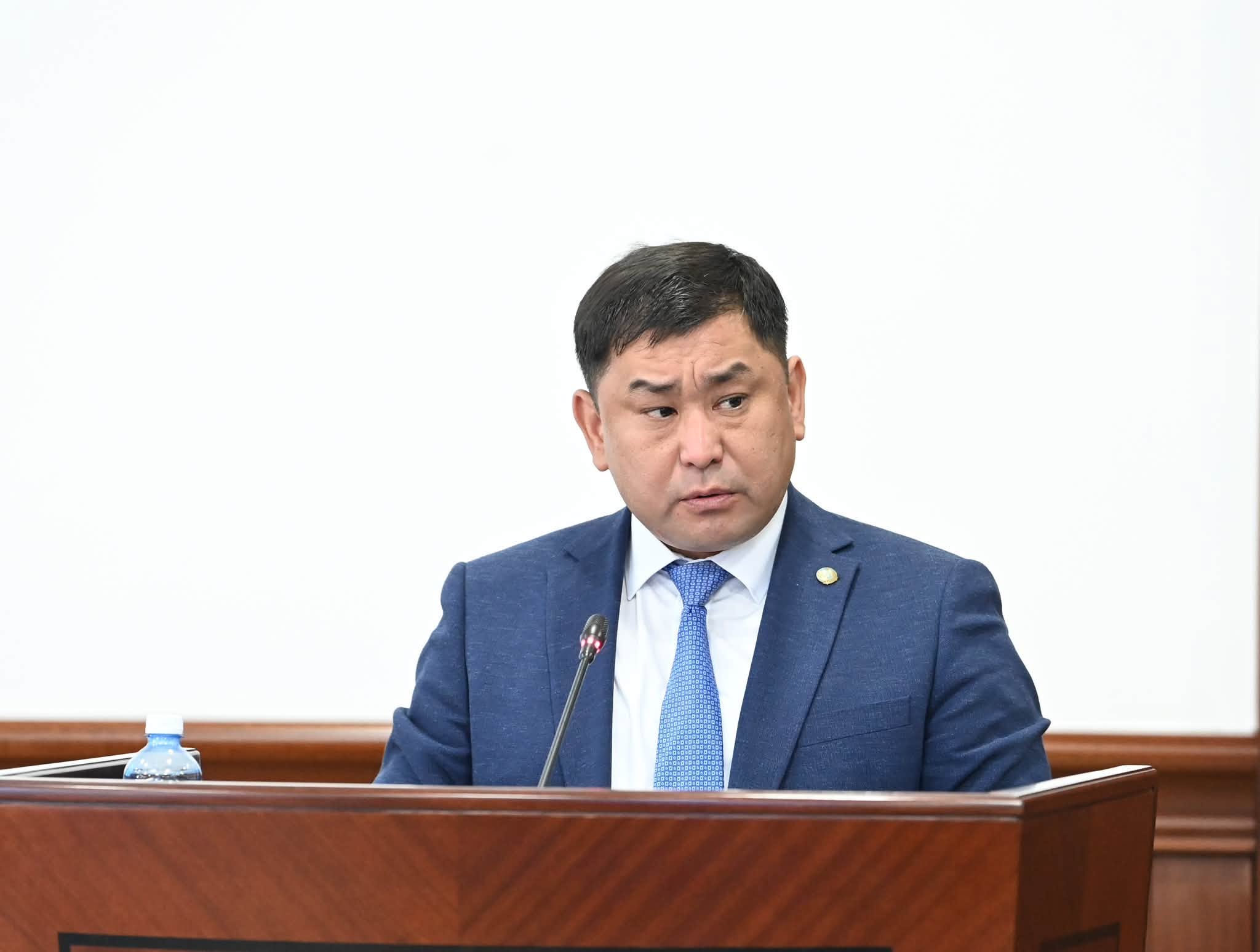 Көк костюм мен галстук киген ер адам трибунада сөйлеп тұр.