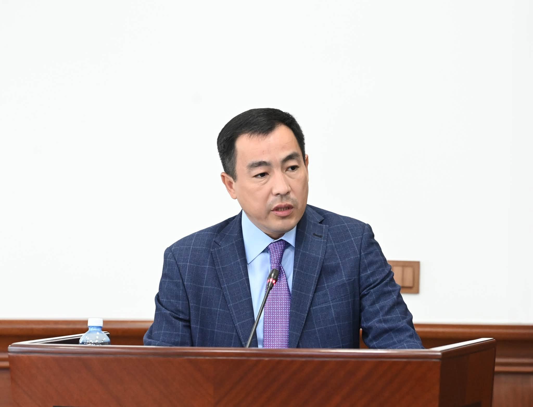 Көк костюм мен күлгін галстук киген ер адам трибунада сөйлеп тұр.