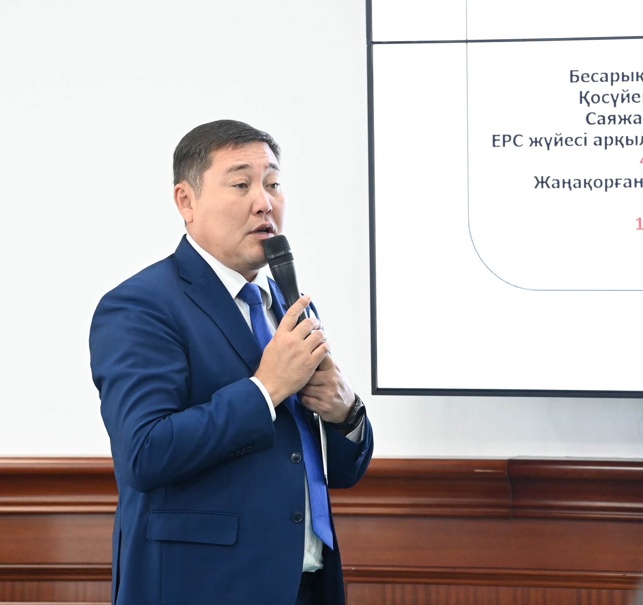 Көк костюм киген ер адам микрофонмен баяндама жасап тұр.
