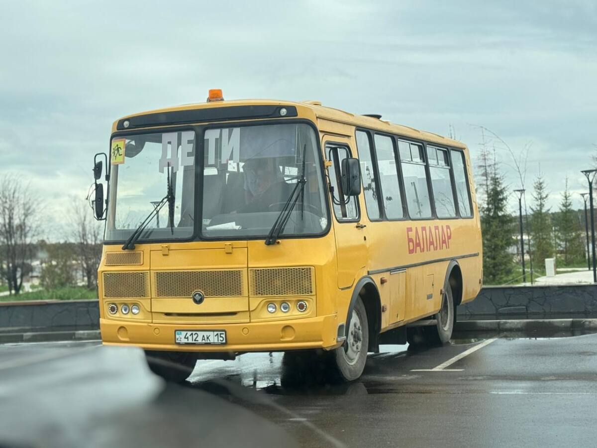 Желтый школьный автобус с надписью «ДЕТИ» на лобовом стекле стоит на мокрой парковке в пасмурный день.