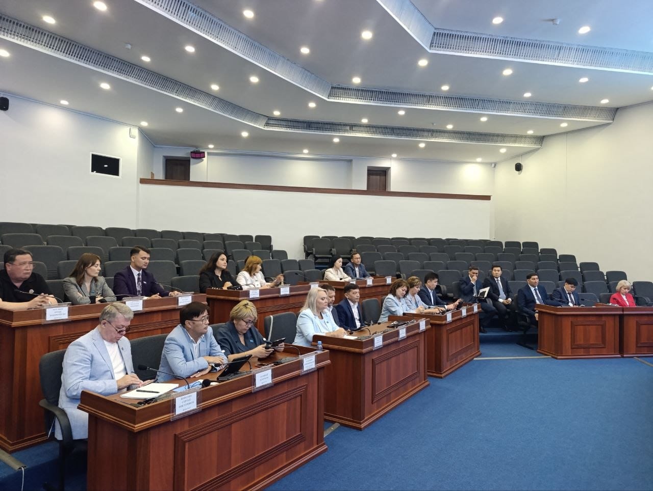 Іскерлік киім киген бір топ адам ресми іс-шара кезінде конференц-залдағы үстелдерде отырады.