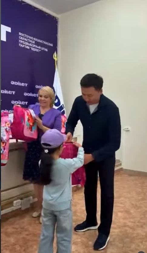 "Әділет"партиясының баннері аясында кішкентай қызға мектеп керек-жарақтары бар рюкзактарды тапсыру.