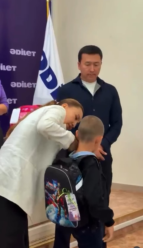 "Әділет"партиясының баннері аясында кішкентай балаға мектеп керек-жарақтары бар рюкзактарды тапсыру.